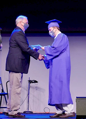 Young man in a graduation cap and gown receiving a diploma from his teacher